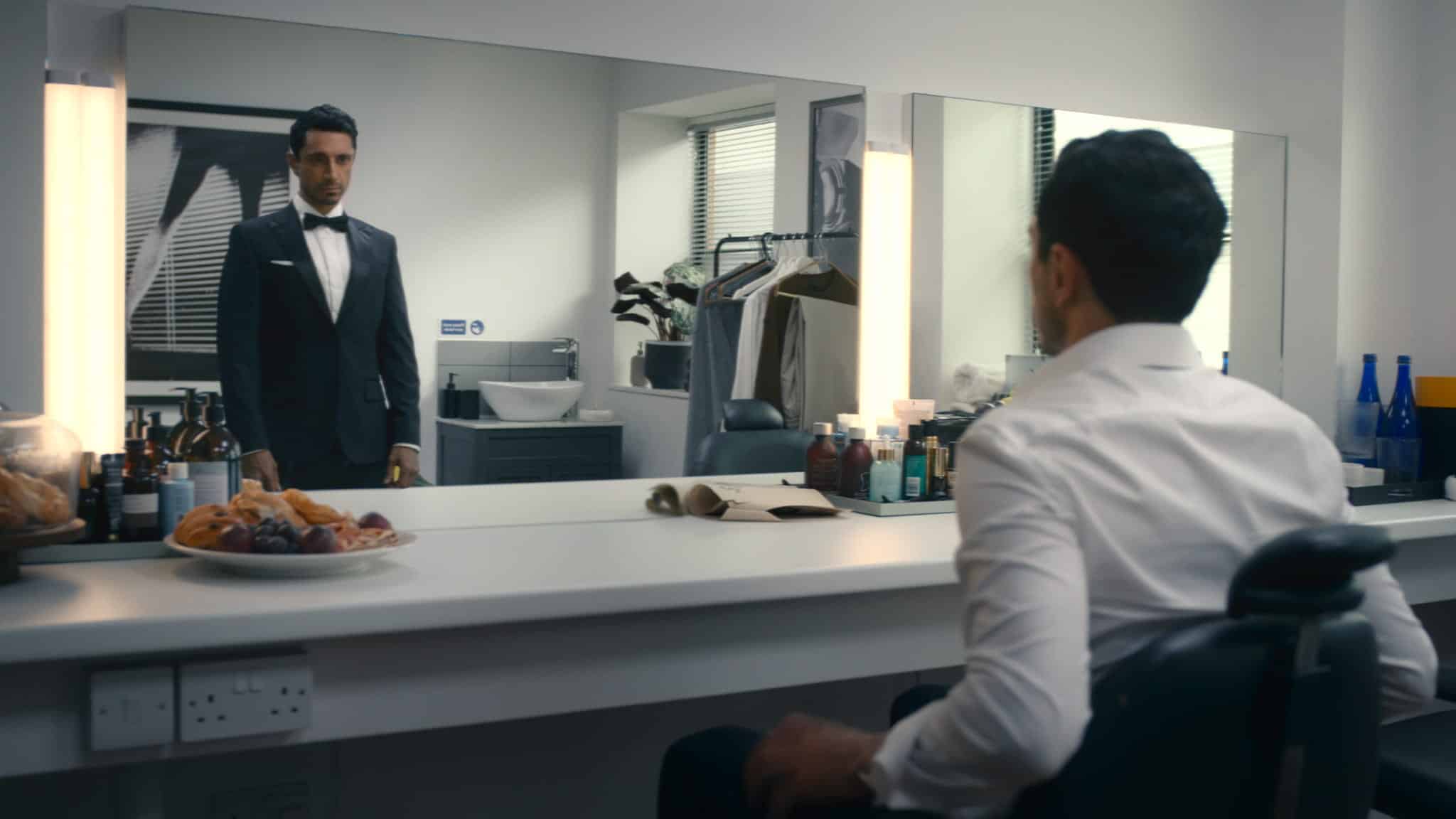 A man in a dressing room sits at a makeup counter, wearing a white shirt and staring at his reflection in the mirror, where he appears dressed in a black tuxedo. Toiletries and snacks are on the counter.