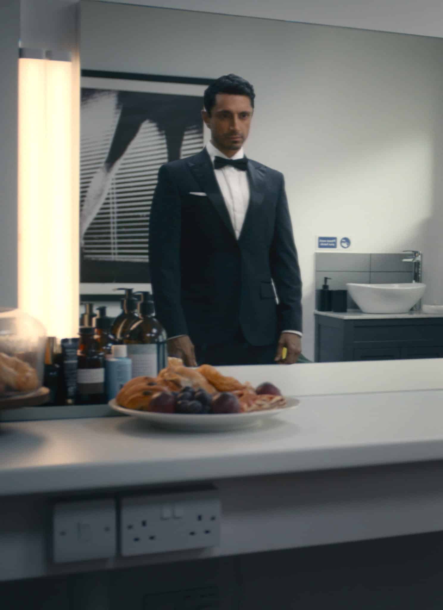 A man in a dressing room sits at a makeup counter, wearing a white shirt and staring at his reflection in the mirror, where he appears dressed in a black tuxedo. Toiletries and snacks are on the counter.