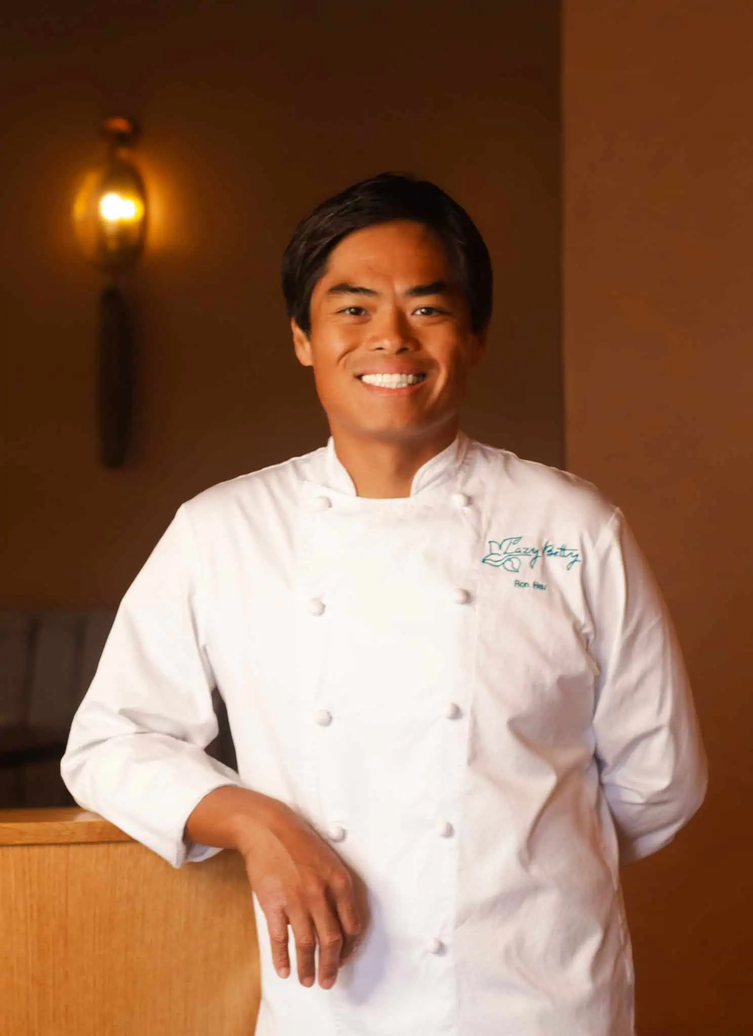 A smiling chef, Ron Hsu, in a white coat stands next to the cover of the cookbook Down South + East: A Chinese American Cookbook, which shows fried chicken being drizzled with honey on a plate.