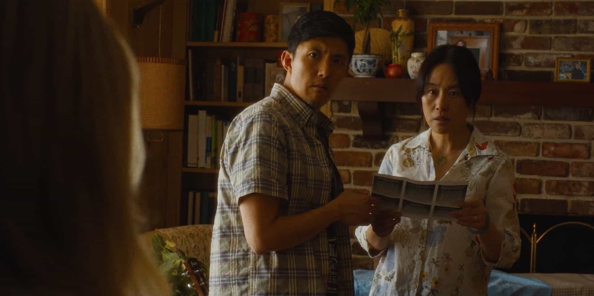 A man and woman stand indoors, looking concerned while holding papers. The background shows a brick wall with shelves of books and decorative items. Warm lighting creates a tense, dramatic atmosphere.