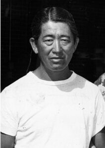 Black and white photo of Soichi Sakamoto, a man with dark, slicked-back hair, wearing a plain white T-shirt, looking at the camera with a neutral expression against a dark background.