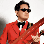 A man in a bright red suit with a black bow tie and dark sunglasses holds an orange electric guitar, standing against a plain white background.
