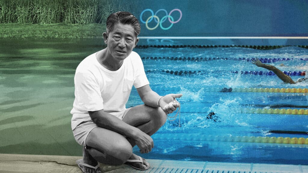 A man in a white t-shirt and shorts squats by a swimming pool, holding a stopwatch. Swimmers compete in lanes behind him, and Olympic rings are visible on a blue background above the pool.