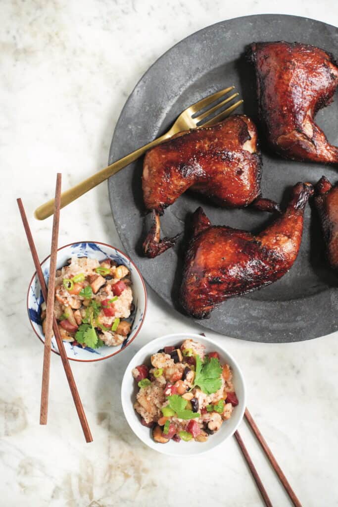 A dark plate with three pieces of glazed roasted chicken, a gold fork, and two bowls of mixed bean salad with cilantro, alongside wooden chopsticks on a light marble surface.