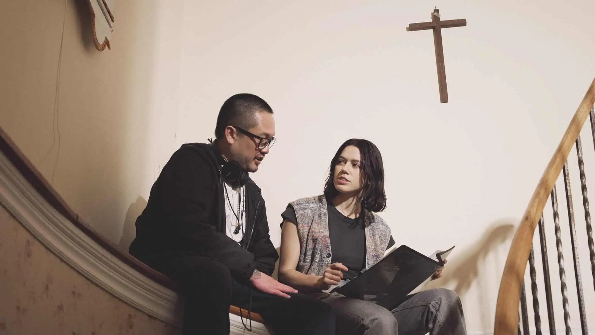 Two people sit on a curved stair landing, talking. One holds a notebook, the other has headphones around their neck. A large wooden cross hangs on the wall above them. The setting appears to be indoors, near a staircase.