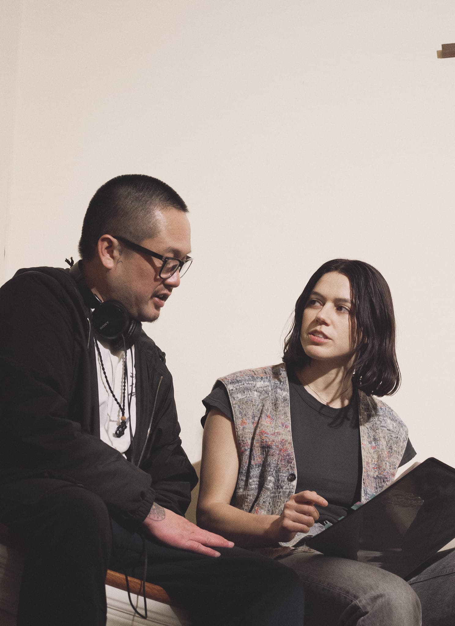 Two people sit on a curved stair landing, talking. One holds a notebook, the other has headphones around their neck. A large wooden cross hangs on the wall above them. The setting appears to be indoors, near a staircase.