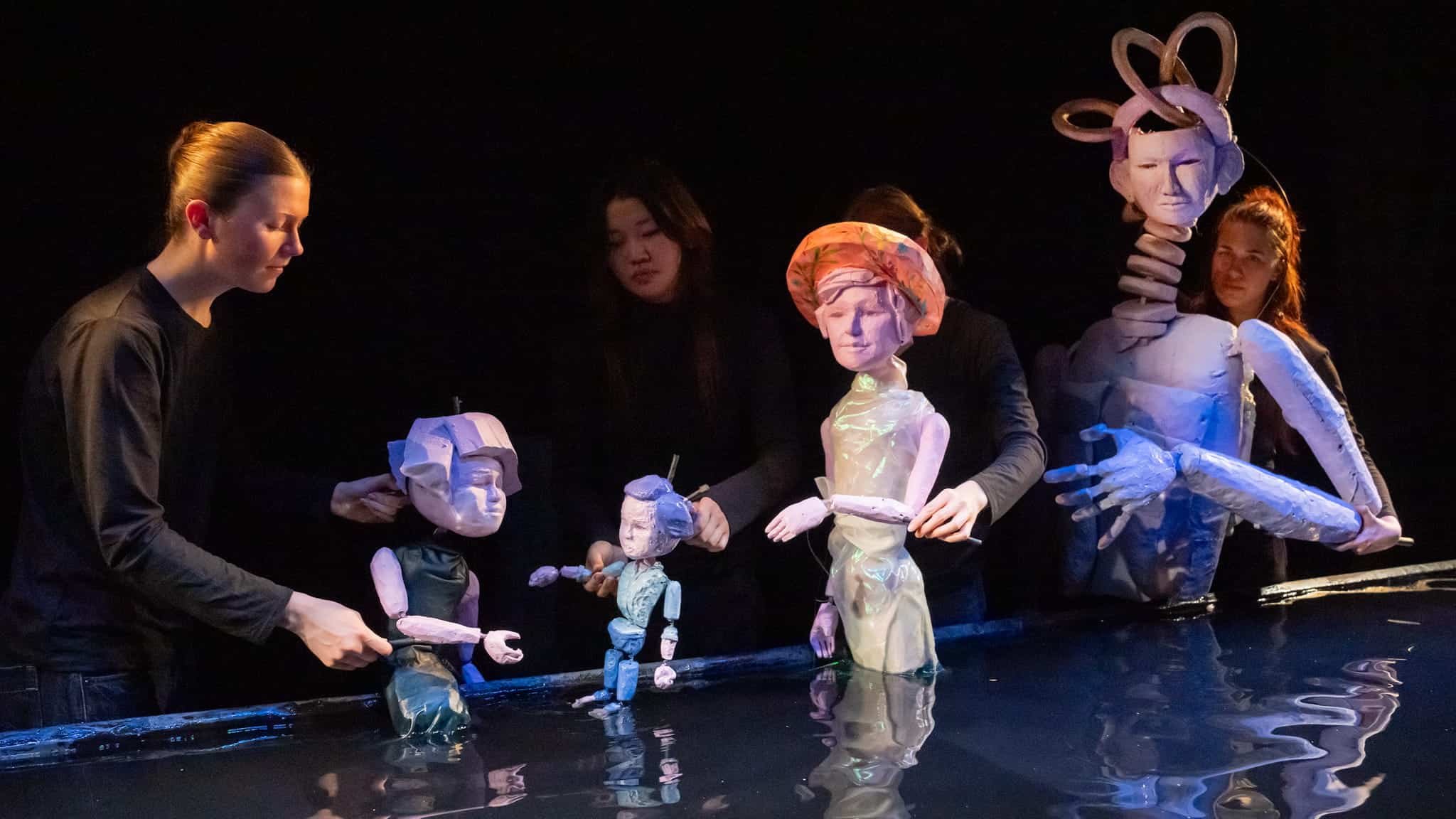 Five puppeteers dressed in black manipulate various large, artistic puppets with human-like features and creative designs on a dark stage, with reflected light on a shiny surface below.
