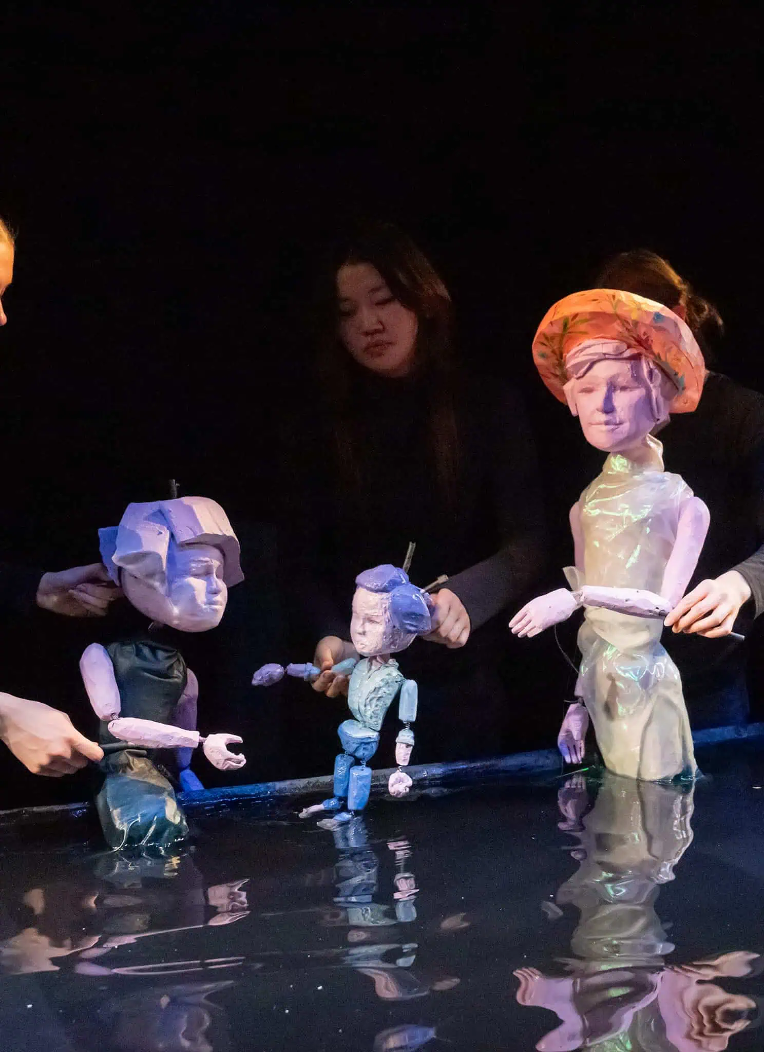 Five puppeteers dressed in black manipulate various large, artistic puppets with human-like features and creative designs on a dark stage, with reflected light on a shiny surface below.