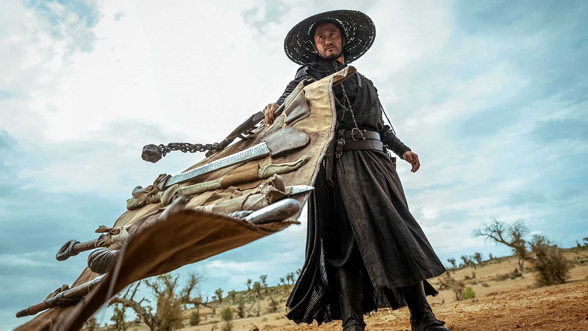 A man dressed in black robes and a wide-brimmed hat stands on arid ground, holding a large, rolled-up weapon or bundle with multiple swords and chains, looking intense under a cloudy sky.