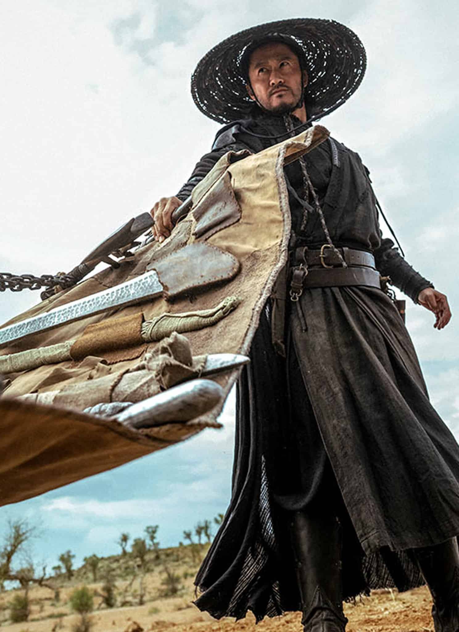 A man dressed in black robes and a wide-brimmed hat stands on arid ground, holding a large, rolled-up weapon or bundle with multiple swords and chains, looking intense under a cloudy sky.