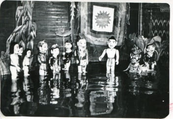 Black and white photo of traditional puppets standing in water, with a tapestry in the background featuring a sun design. The scene appears to be part of a puppet show, staged with detailed props and backdrop.