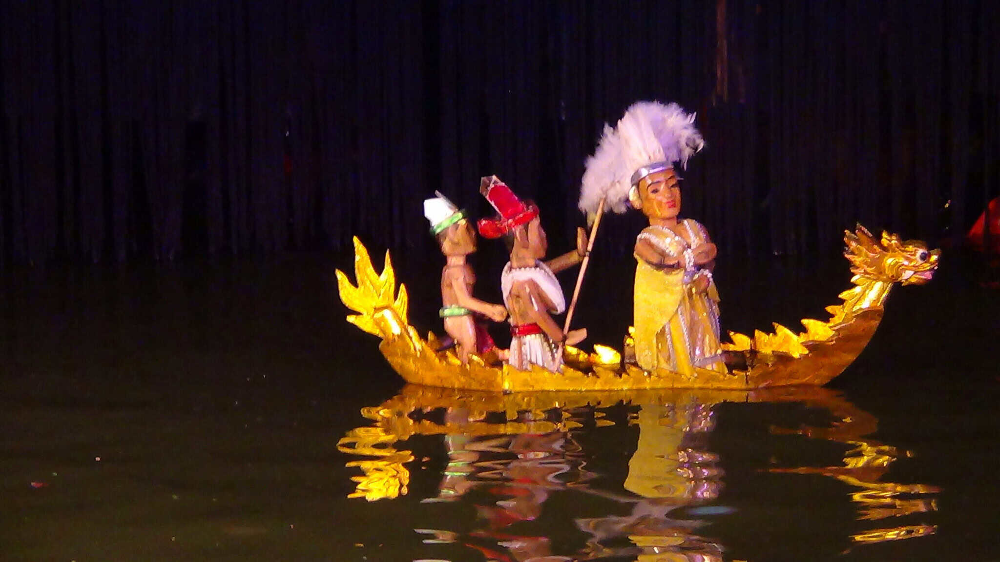 Three colorful puppets on a golden dragon boat float on water during a traditional Vietnamese water puppet show, with dark curtains in the background and reflections visible on the water.
