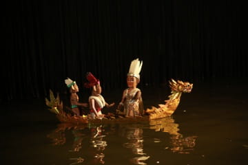 Three puppets dressed in traditional attire sit in a decorative golden dragon boat, floating on water against a dark background.