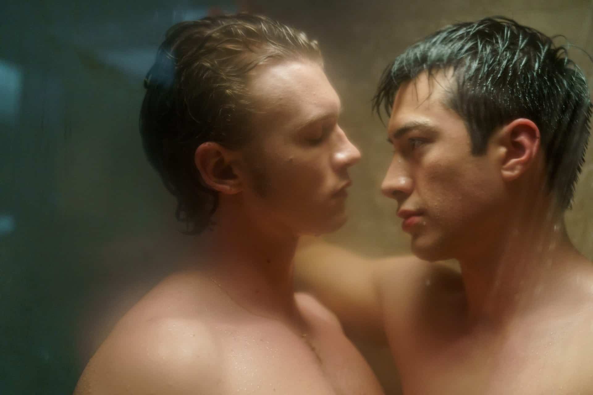 Two shirtless men stand close together in a steamy, dimly lit setting, gazing intently into each others eyes, suggesting an intimate or emotional moment.