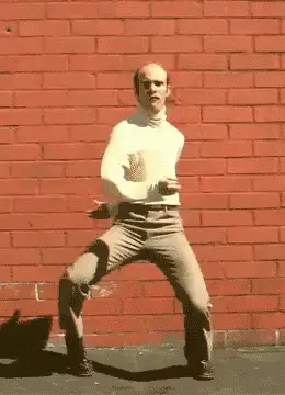 A man with a bald head wearing a white turtleneck and beige pants energetically dances in front of a red brick wall.