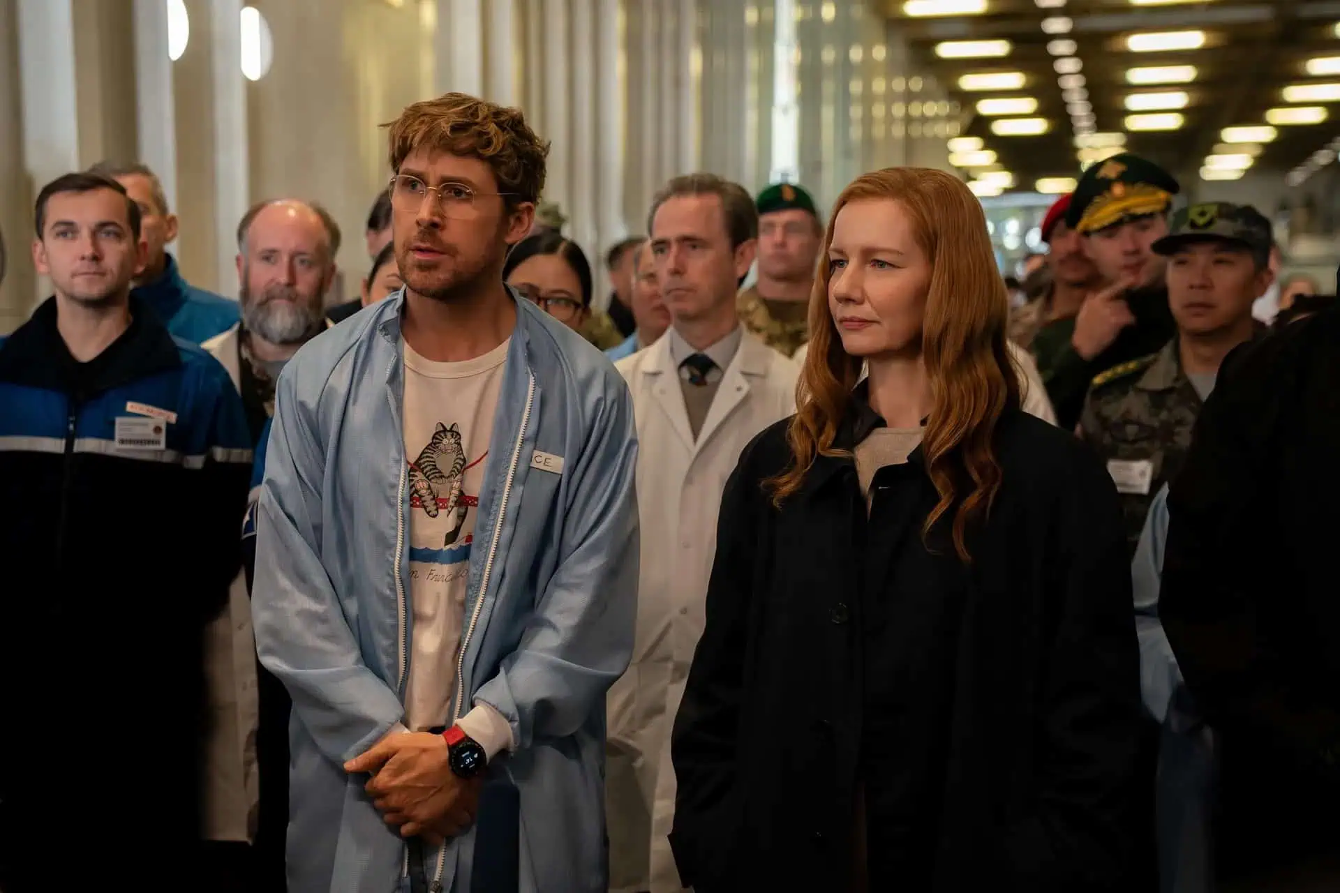A group of people, including scientists in lab coats and individuals in uniforms, stand closely together indoors, listening attentively. Two people, a man in a blue jacket and a woman in dark clothing, are prominent in the foreground.