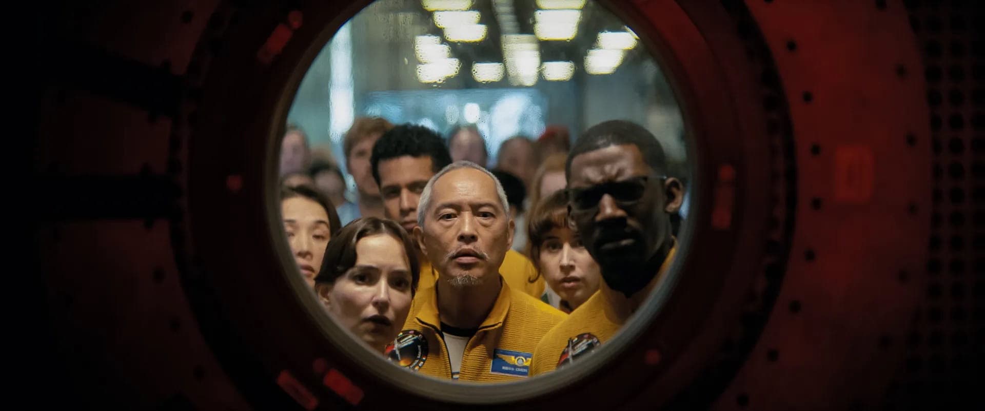 A group of astronauts in yellow suits closely peer through a circular window or hatch, looking serious and curious, with the background blurred and industrial lights above.