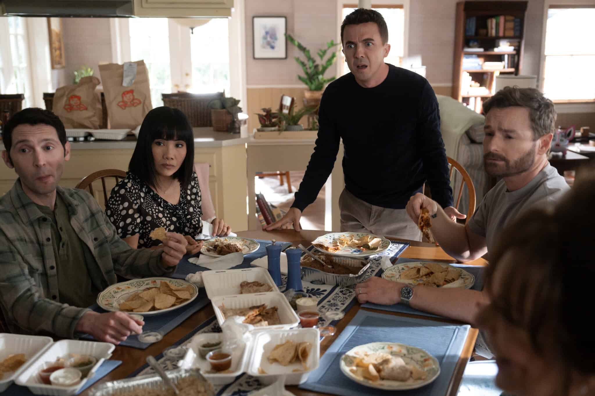 Four people sit at a kitchen table eating takeout food, while one person stands looking surprised. The dining area is bright with grocery bags in the background and food spread out on the table.