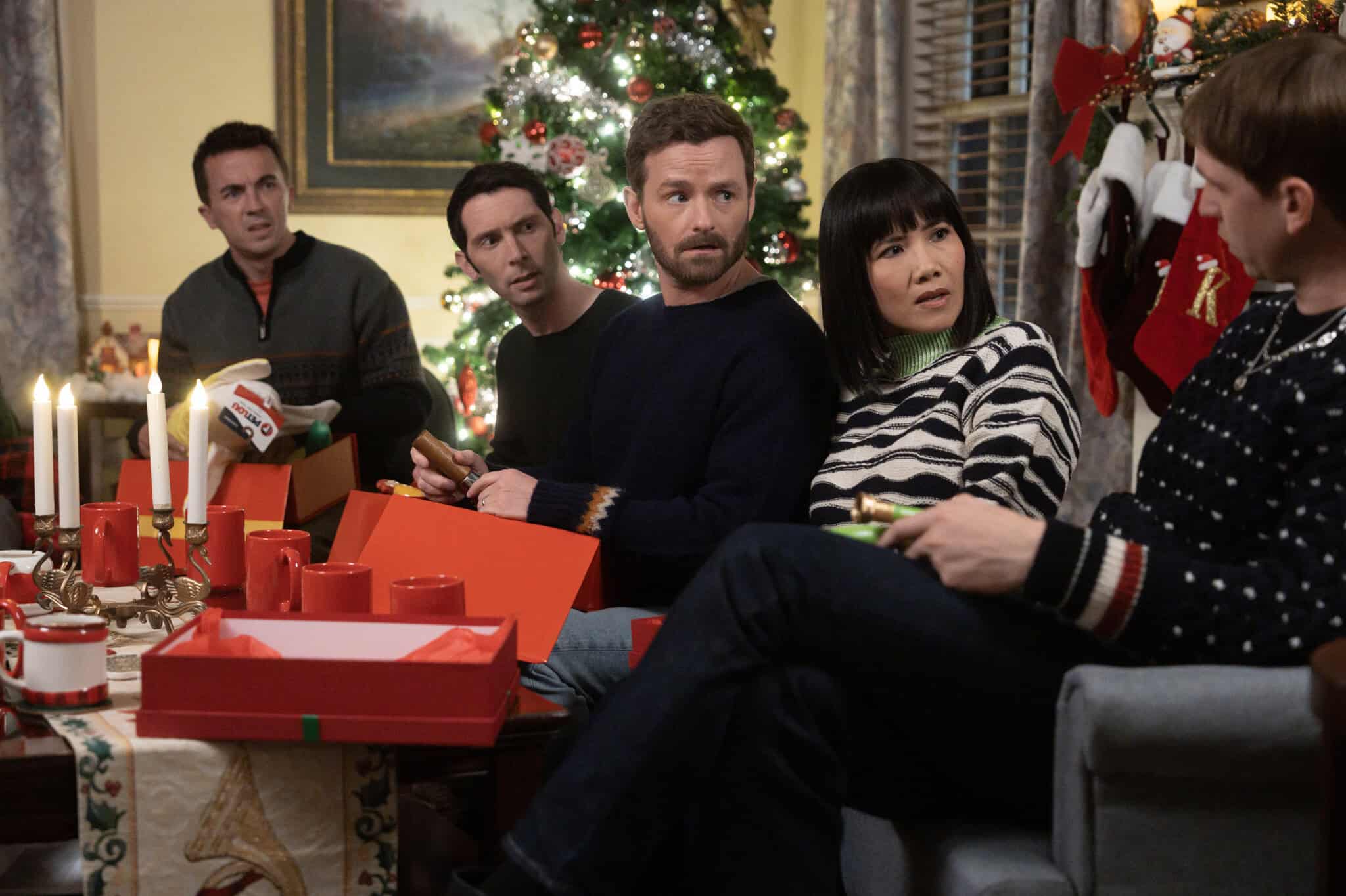 Five people sit around a coffee table with candles and gifts, looking surprised or concerned. Behind them is a decorated Christmas tree and stockings hanging by a window, creating a festive holiday atmosphere.