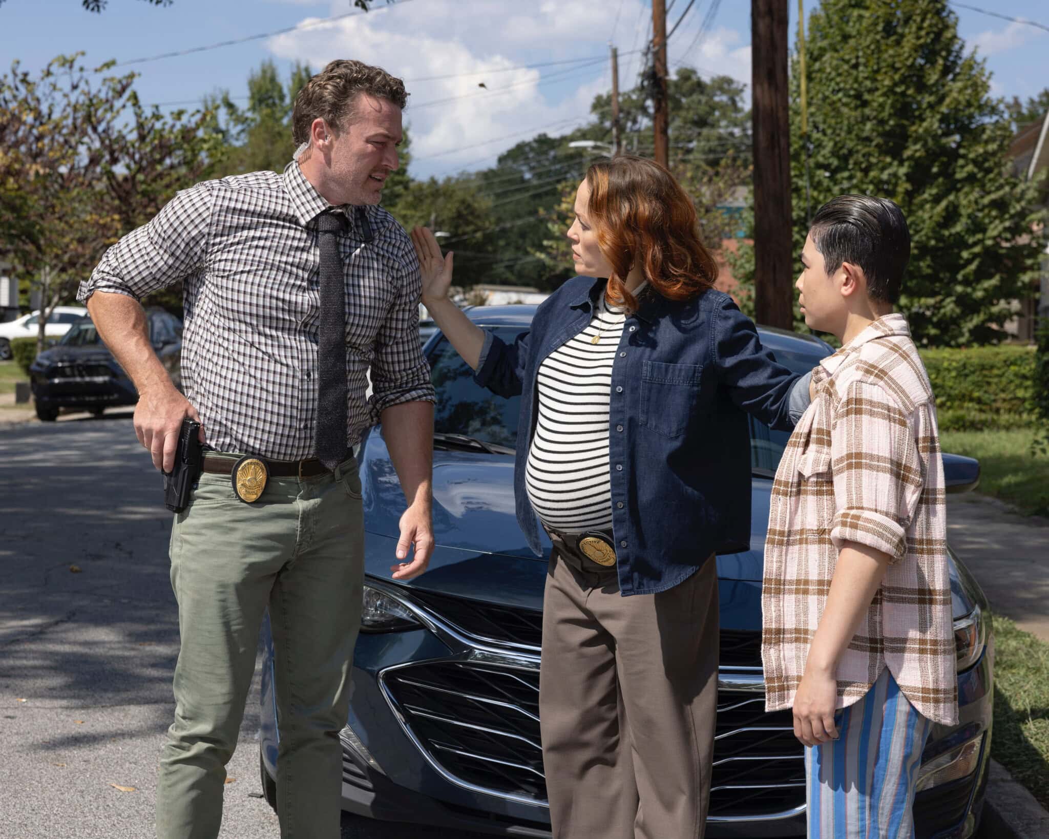Three detectives, including Cora Lu and Will Trent, stand in front of a car on a sunny suburban street. One is visibly pregnant and gesturing while talking to the others, who listen attentively with their badges visible.