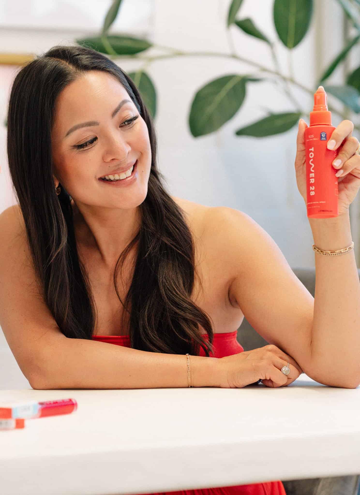 A woman sitting at a table smiles and holds up an orange skincare product. Several other similar orange skincare products are arranged on the table. A vase of orange flowers and green plants are in the background.