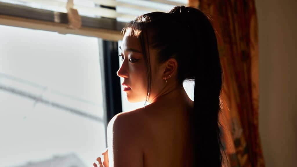A woman with long, dark hair tied in a high ponytail stands near a sunlit window, looking over her shoulder into the light, with shadows falling across her face and back.