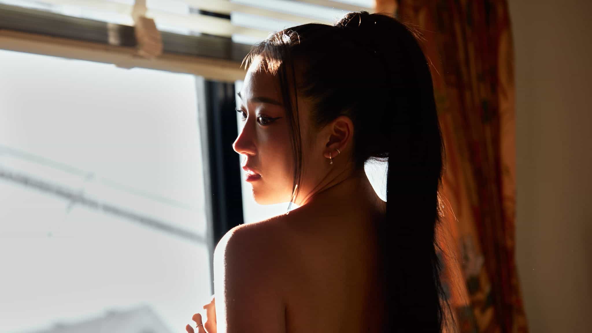 A woman with long, dark hair tied in a high ponytail stands near a sunlit window, looking over her shoulder into the light, with shadows falling across her face and back.