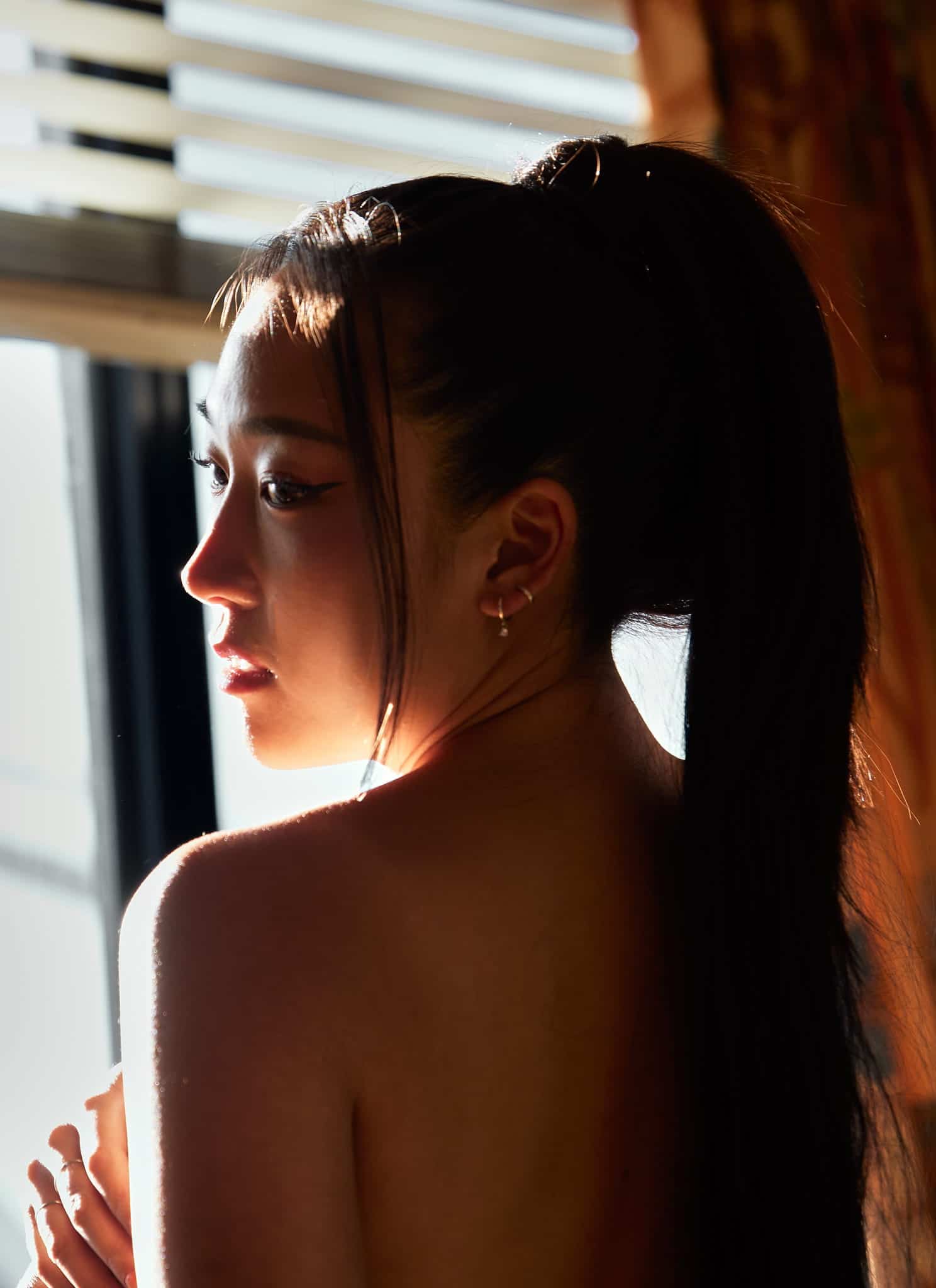 A woman with long, dark hair tied in a high ponytail stands near a sunlit window, looking over her shoulder into the light, with shadows falling across her face and back.