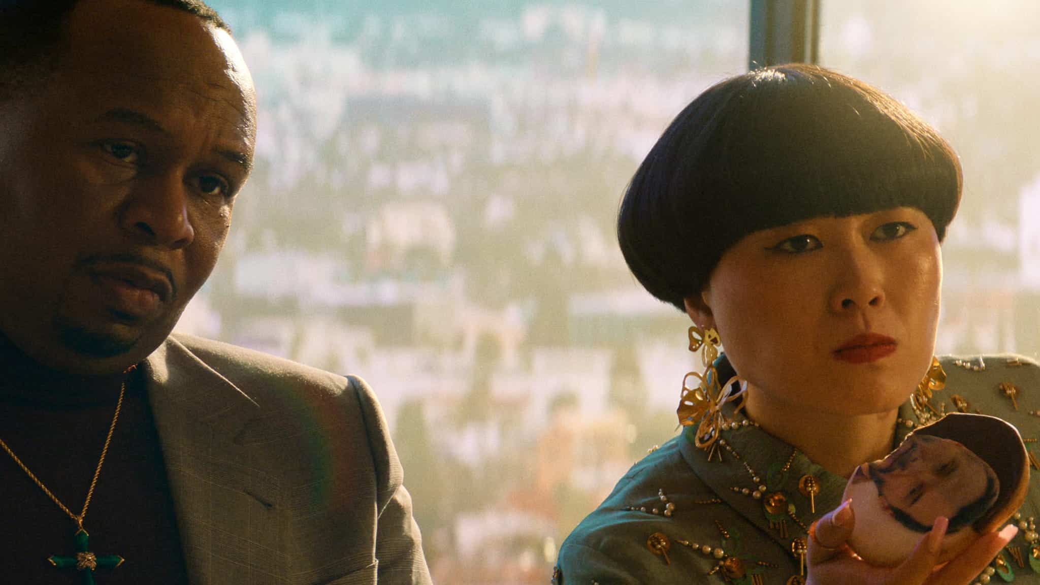 A man in a suit and a woman with a bowl haircut and ornate earrings sit together. The woman holds up a compact mirror showing a reflection of a person’s face. A cityscape is visible through a window behind them.