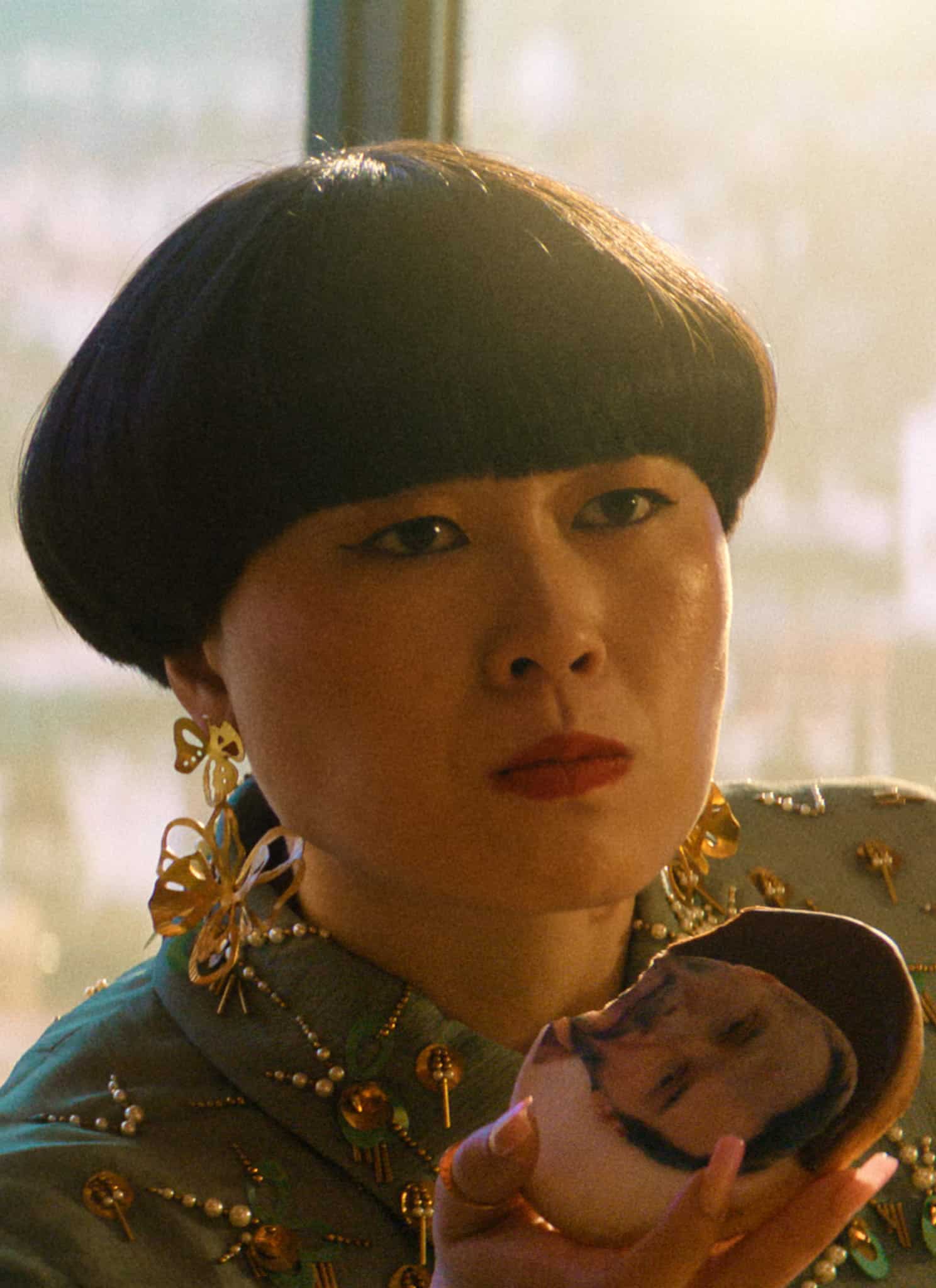 A man in a suit and a woman with a bowl haircut and ornate earrings sit together. The woman holds up a compact mirror showing a reflection of a person’s face. A cityscape is visible through a window behind them.