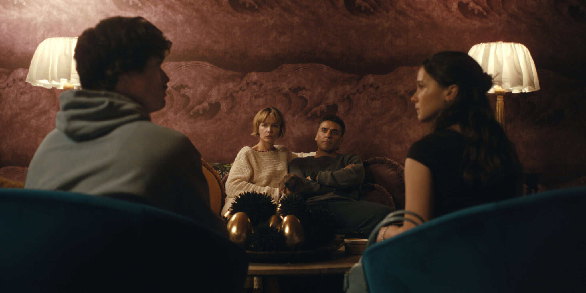Four people sit in a dimly lit room, two facing the camera and two with their backs to it, engaged in a serious conversation around a table with decorative objects. Warm lighting and dark, ornate walls create an intimate atmosphere.