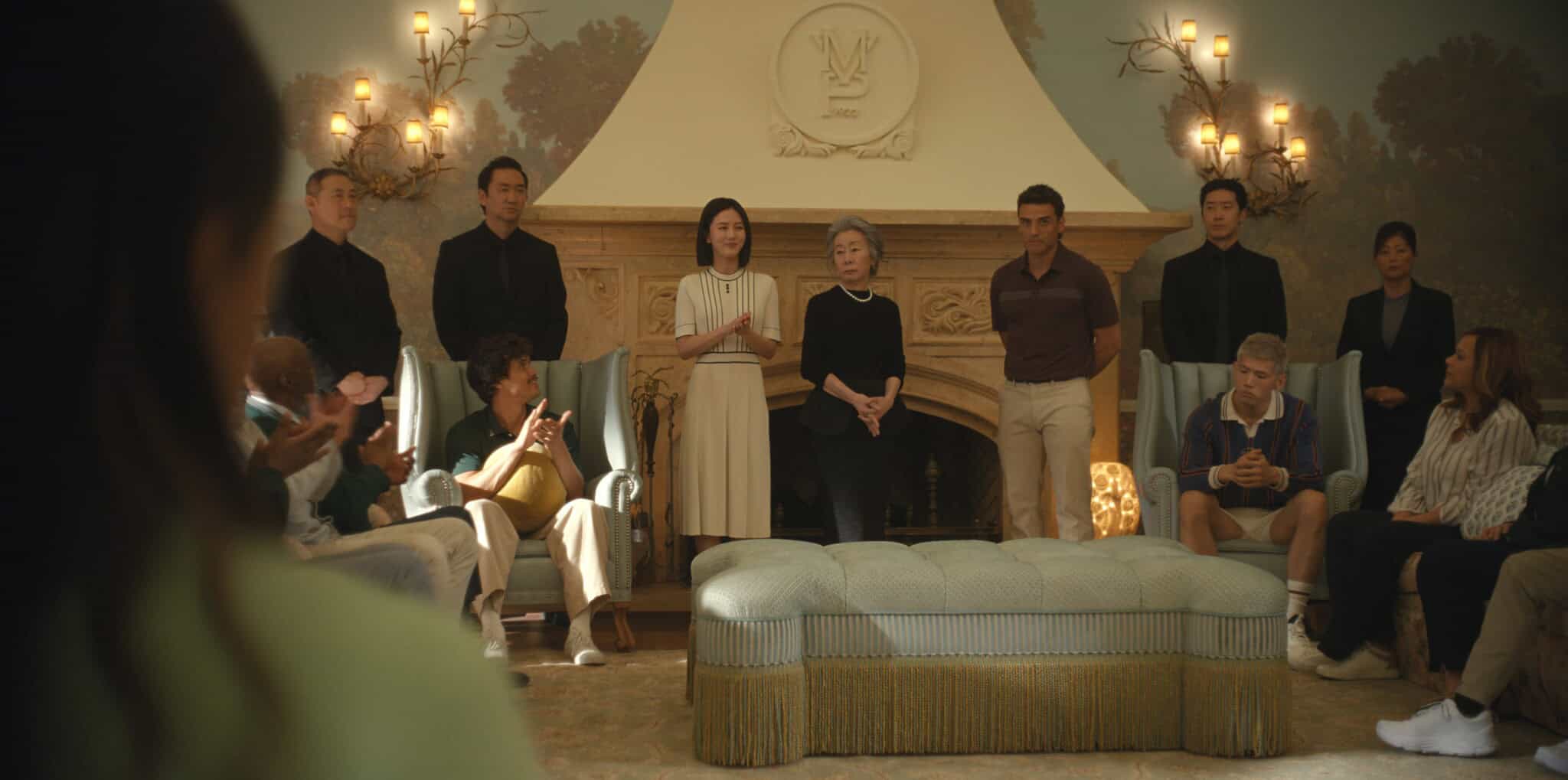 A group of people stand and sit in a luxurious room with elegant lighting and a fireplace. Some are standing in a line facing a seated audience, who are clapping and listening attentively.