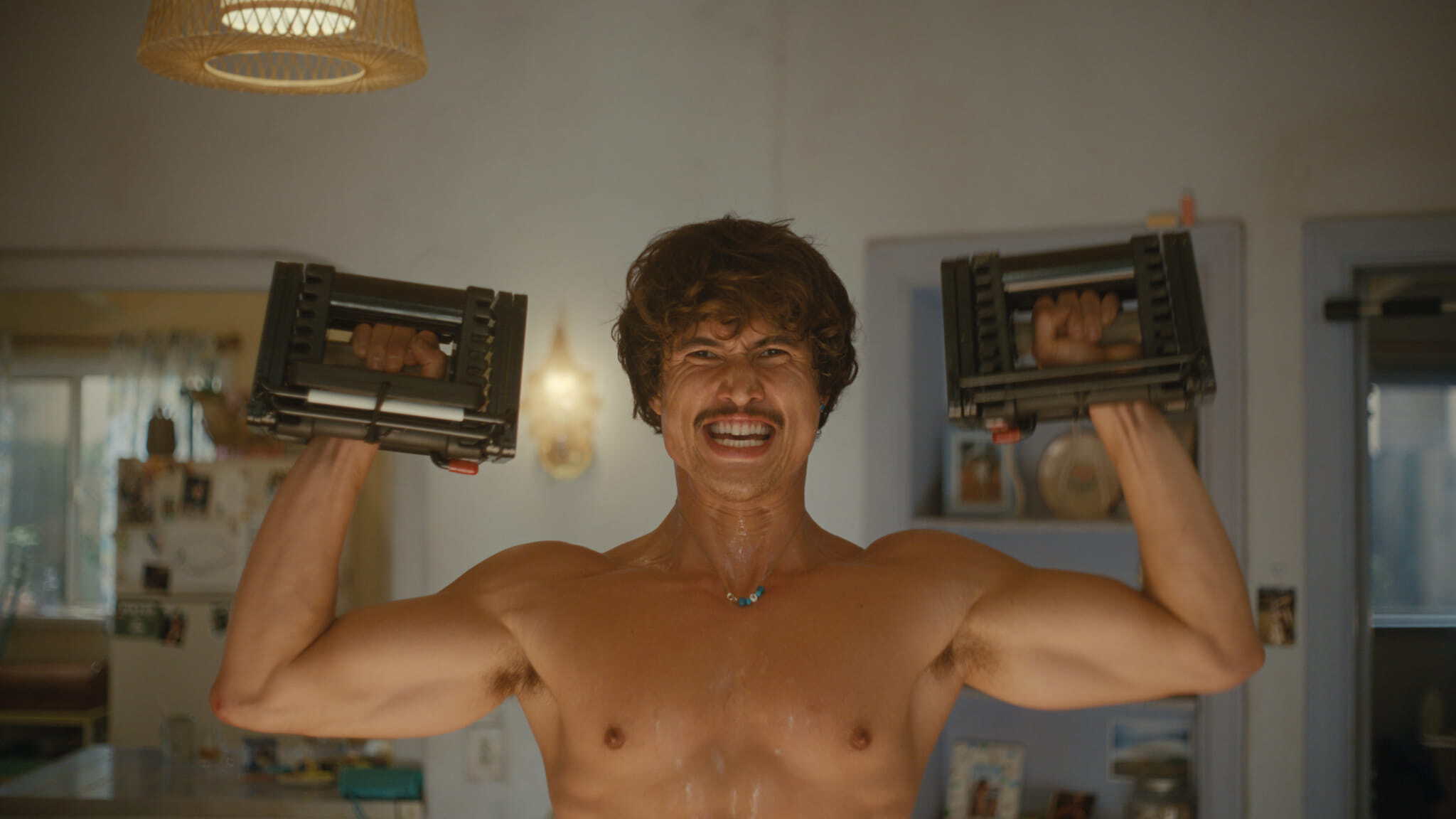 A shirtless man with a mustache and sweaty skin smiles while lifting two adjustable dumbbells above his shoulders in a home setting. The background shows a kitchen area with shelves and hanging lights.