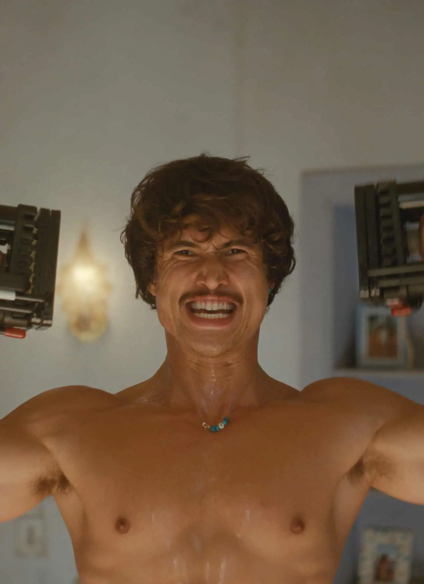 A shirtless man with a mustache and sweaty skin smiles while lifting two adjustable dumbbells above his shoulders in a home setting. The background shows a kitchen area with shelves and hanging lights.