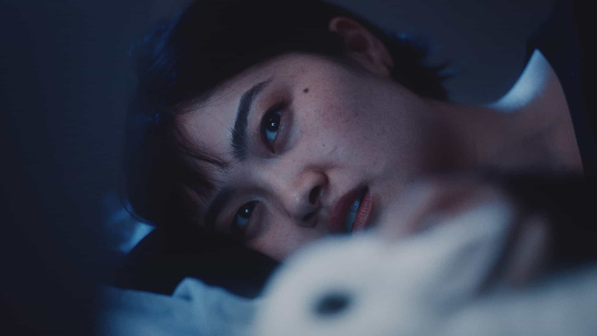 A person with short dark hair lies on their side in dim, blue-tinted lighting, gazing thoughtfully into the distance with a neutral expression.