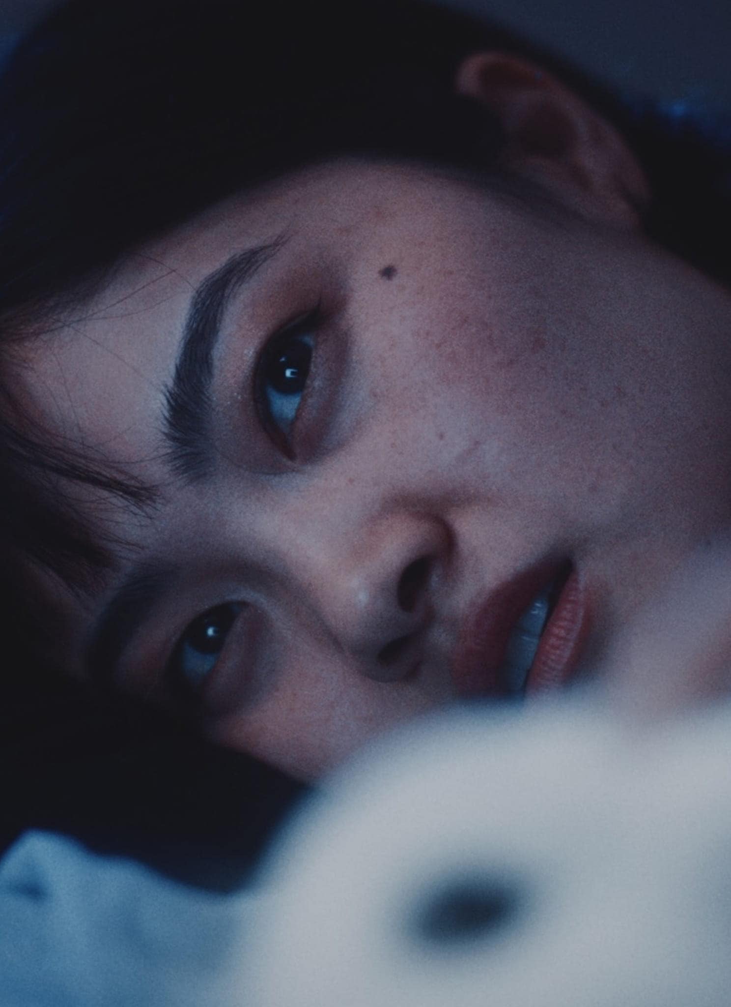 A person with short dark hair lies on their side in dim, blue-tinted lighting, gazing thoughtfully into the distance with a neutral expression.