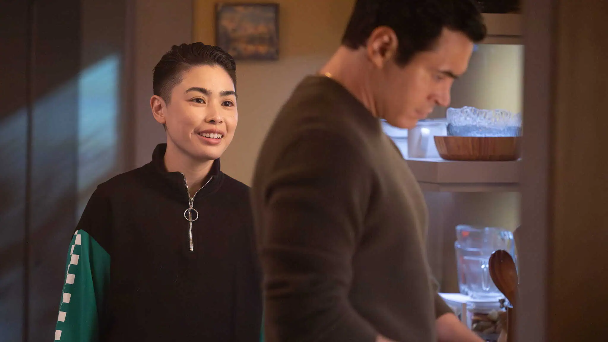 A person with short hair and a dark zip-up jacket smiles while standing behind another person, resembling Cora Lu Tran from Will Trent, who is focused on cooking in a warmly lit kitchen.
