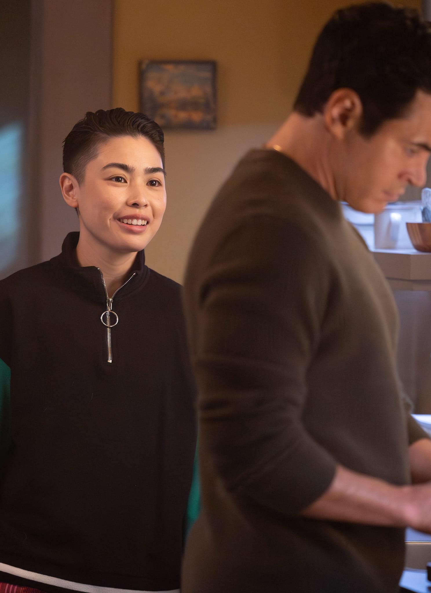 A person with short hair and a dark zip-up jacket smiles while standing behind another person, resembling Cora Lu Tran from Will Trent, who is focused on cooking in a warmly lit kitchen.