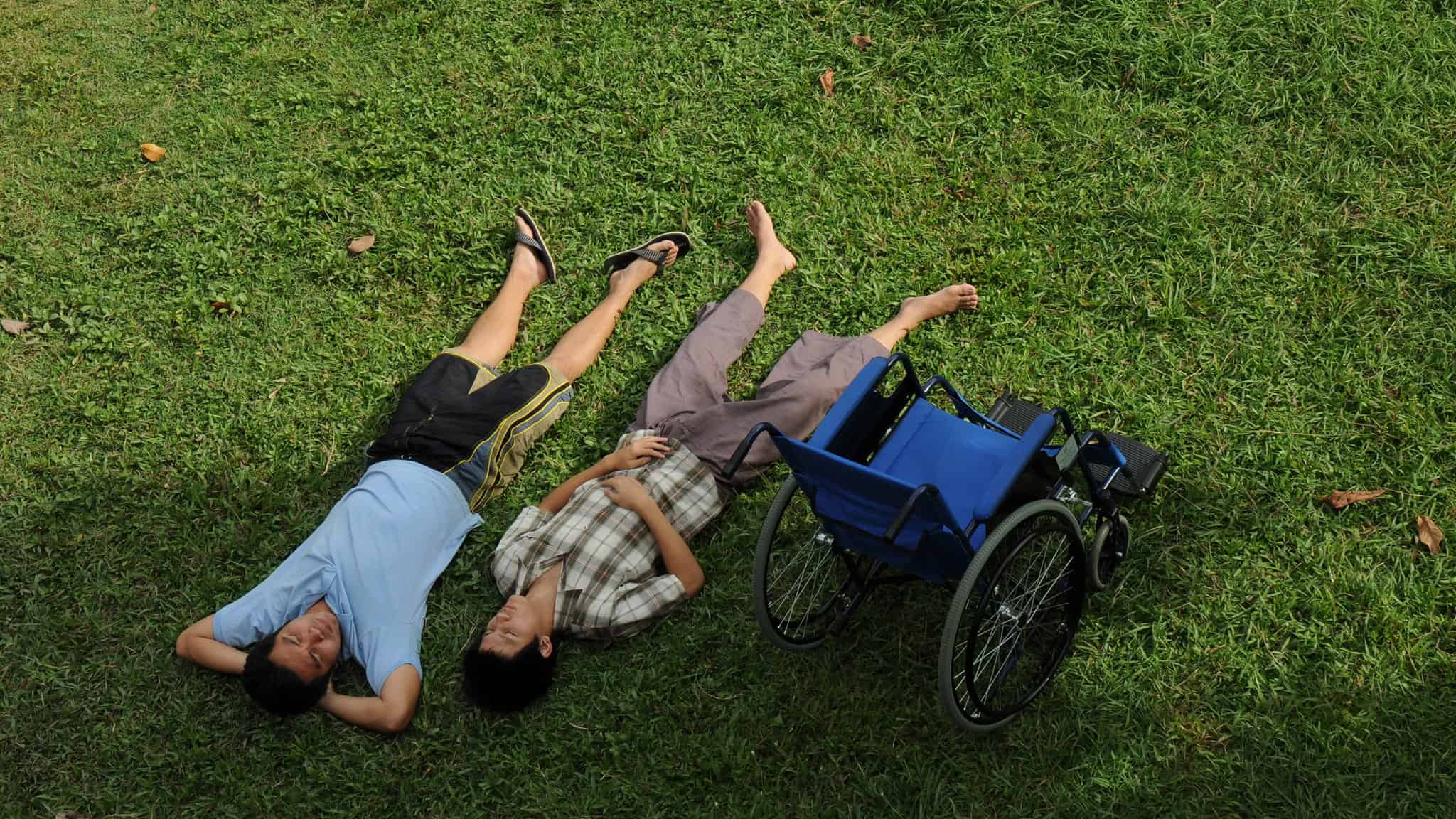 Two people lie side by side on green grass with their legs outstretched, looking up. A blue wheelchair is nearby, unoccupied, indicating relaxation and companionship outdoors.