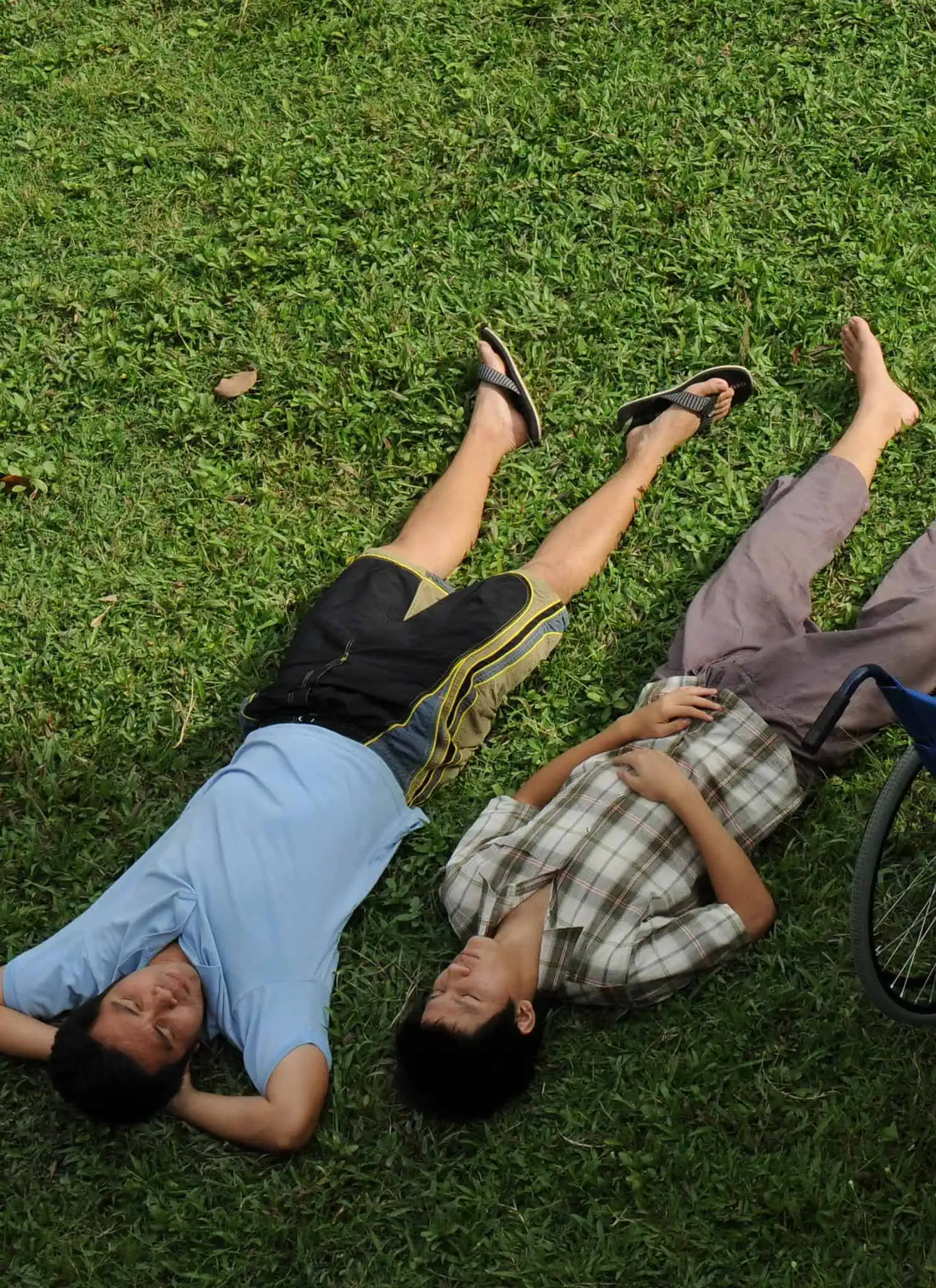 Two people lie side by side on green grass with their legs outstretched, looking up. A blue wheelchair is nearby, unoccupied, indicating relaxation and companionship outdoors.