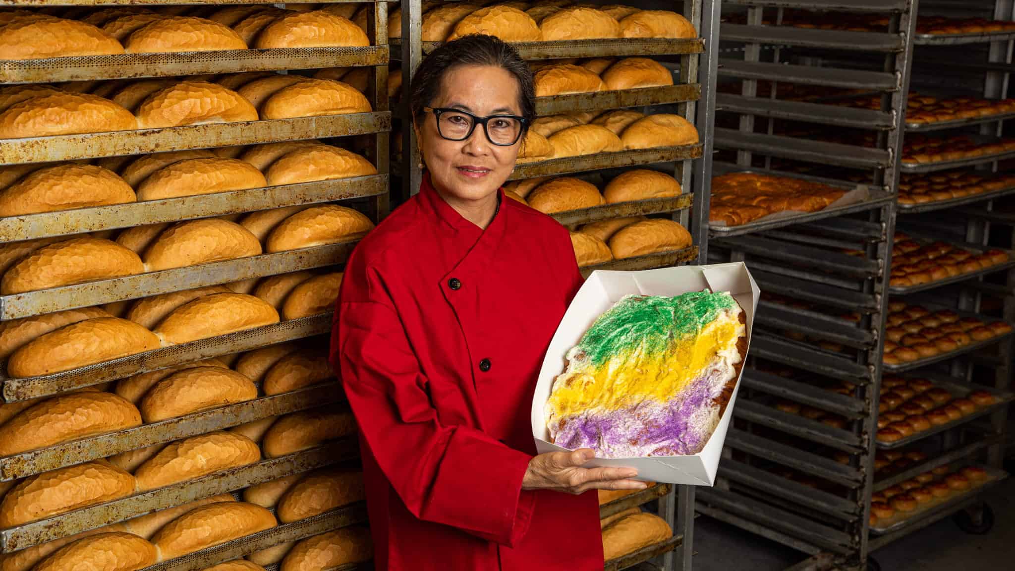 A woman in a red chef’s coat stands in a bakery surrounded by racks of bread, holding a colorful cake with green, yellow, and purple icing in a white box.