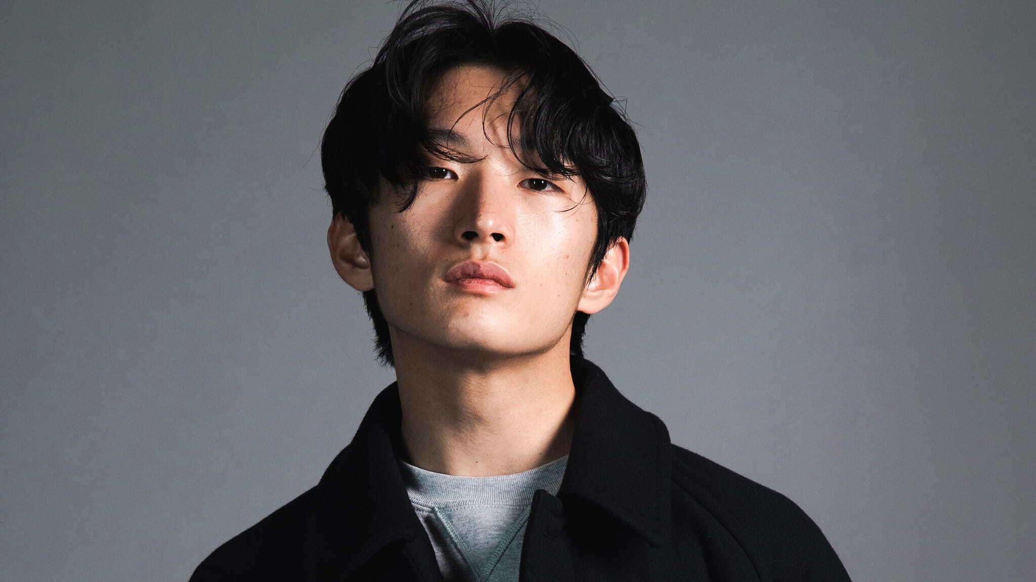 A young man with medium-length black hair and a serious expression looks directly at the camera. He is wearing a dark jacket over a light shirt, with a neutral gray background behind him.
