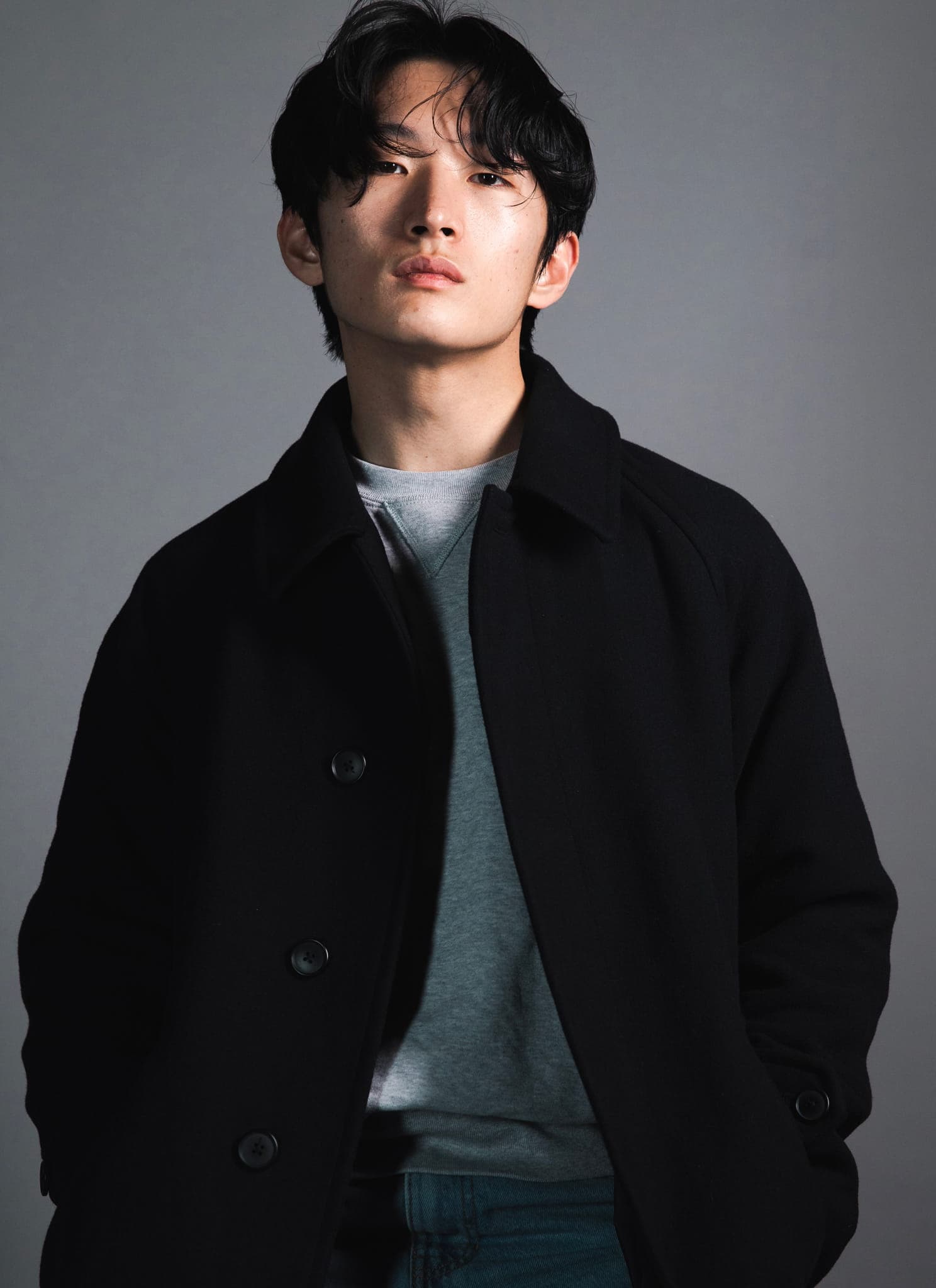A young man with medium-length black hair and a serious expression looks directly at the camera. He is wearing a dark jacket over a light shirt, with a neutral gray background behind him.