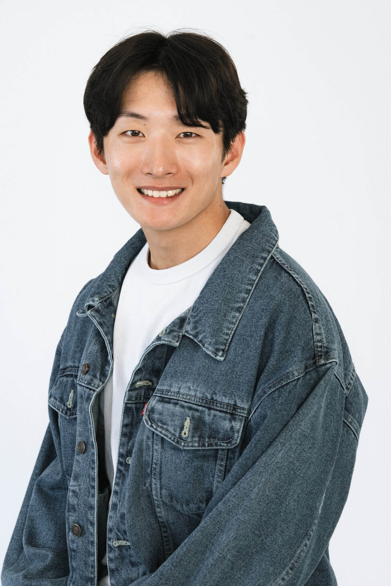 A young person with short black hair smiles at the camera, wearing a white t-shirt and a blue denim jacket. The background is plain white.