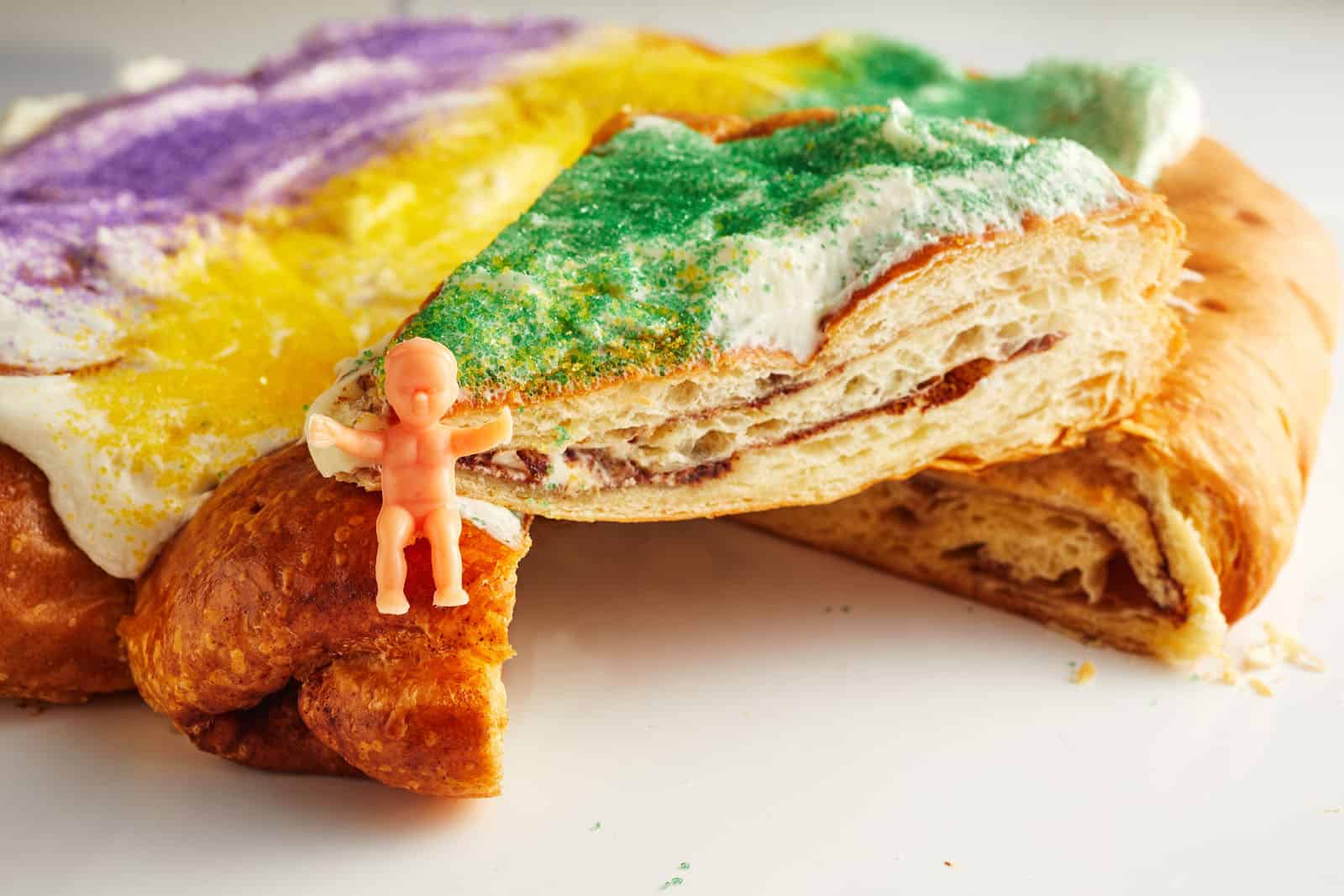 A slice of King Cake topped with yellow, purple, and green sugar sits on a plate, with a small plastic baby figurine placed on the cake.