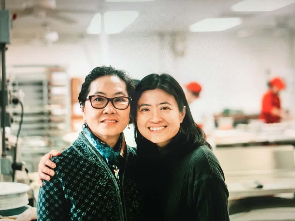 Two women stand close together and smile in a well-lit kitchen or bakery, with shelves and workers in red hats blurred in the background.