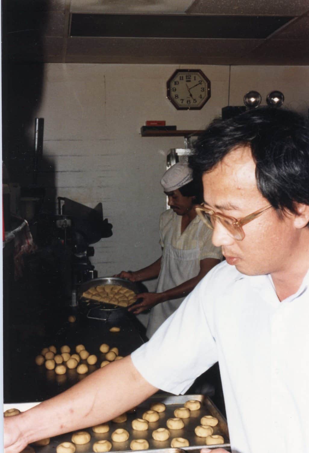 Two men work in a bakery. One in the foreground arranges rows of round pastries on a tray, while another in the background, wearing a bakers hat, stands by trays of pastries. A wall clock shows 7:47.