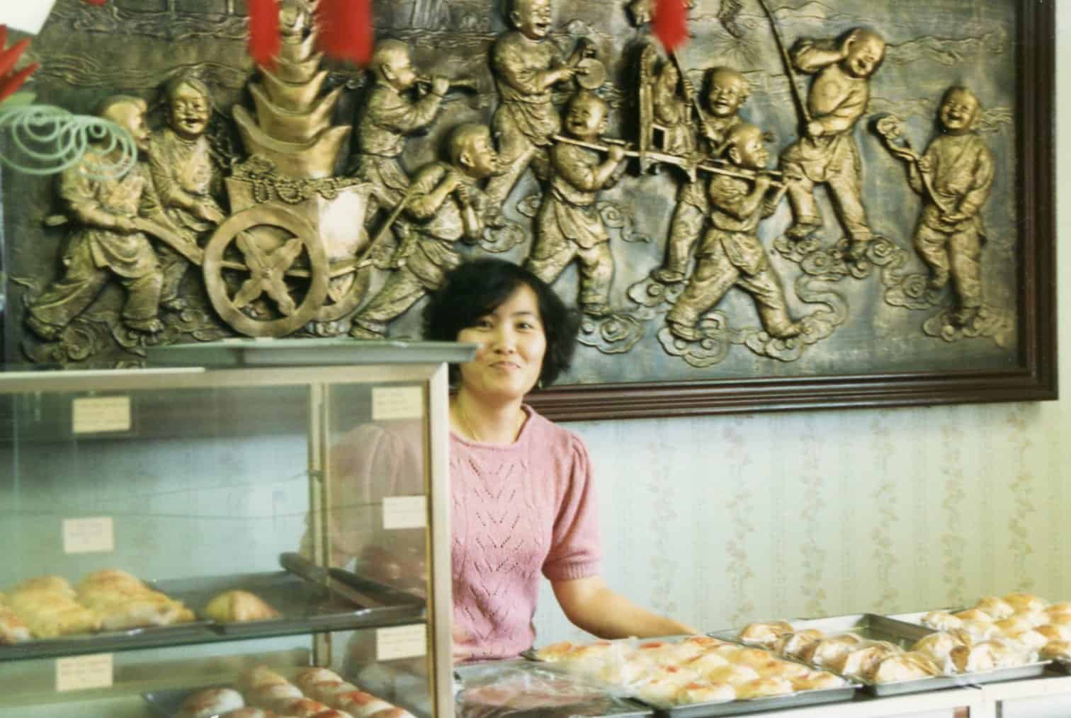 A woman in a pink sweater stands behind a pastry display in a bakery. Behind her is an ornate wall relief depicting a group of people in a festive or historical scene. Pastries are visible in the foreground.