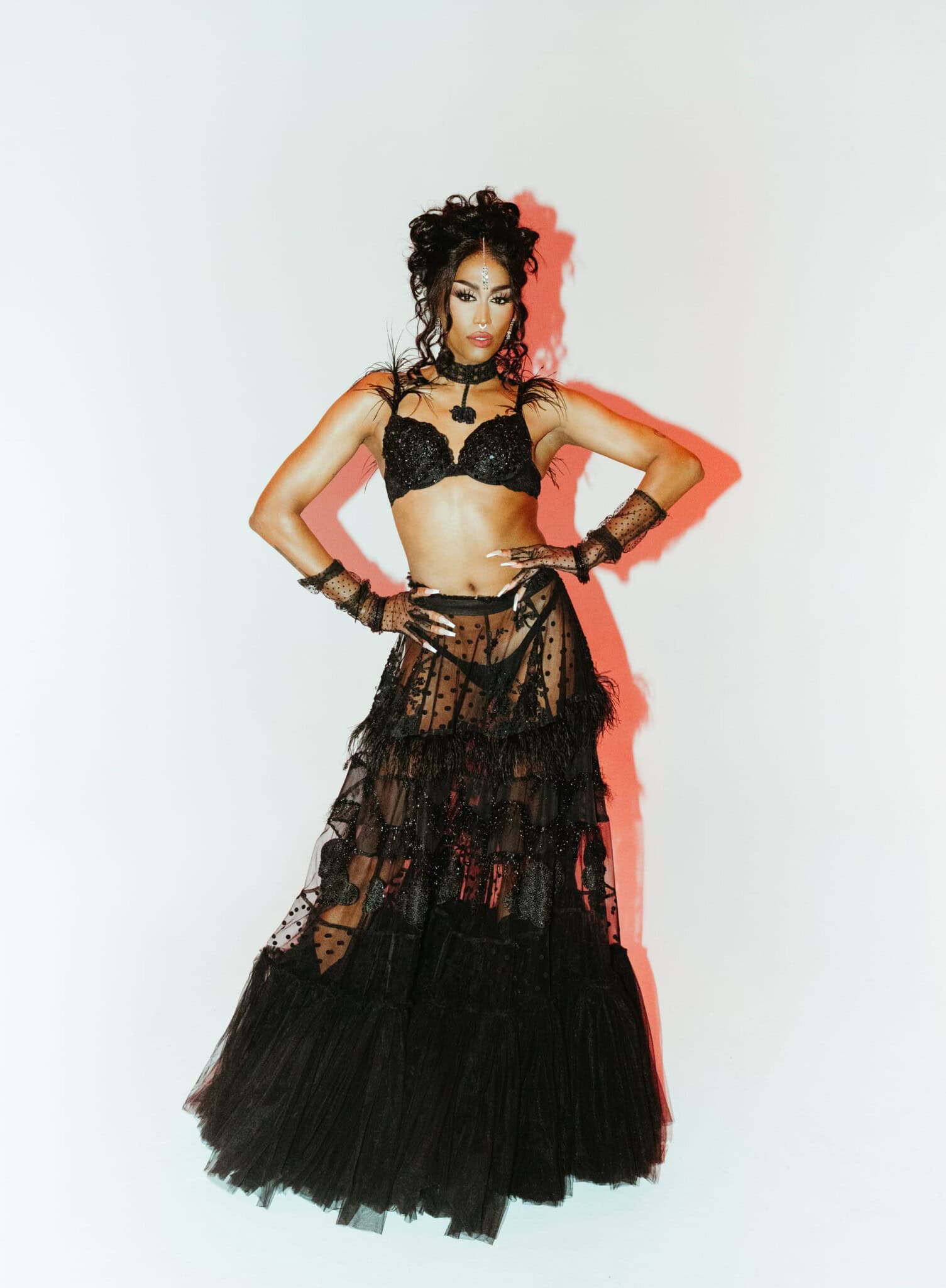 A person poses confidently in a black bralette and a sheer, layered black tulle skirt, accessorized with bracelets, earrings, and a necklace, against a white background with dramatic lighting.