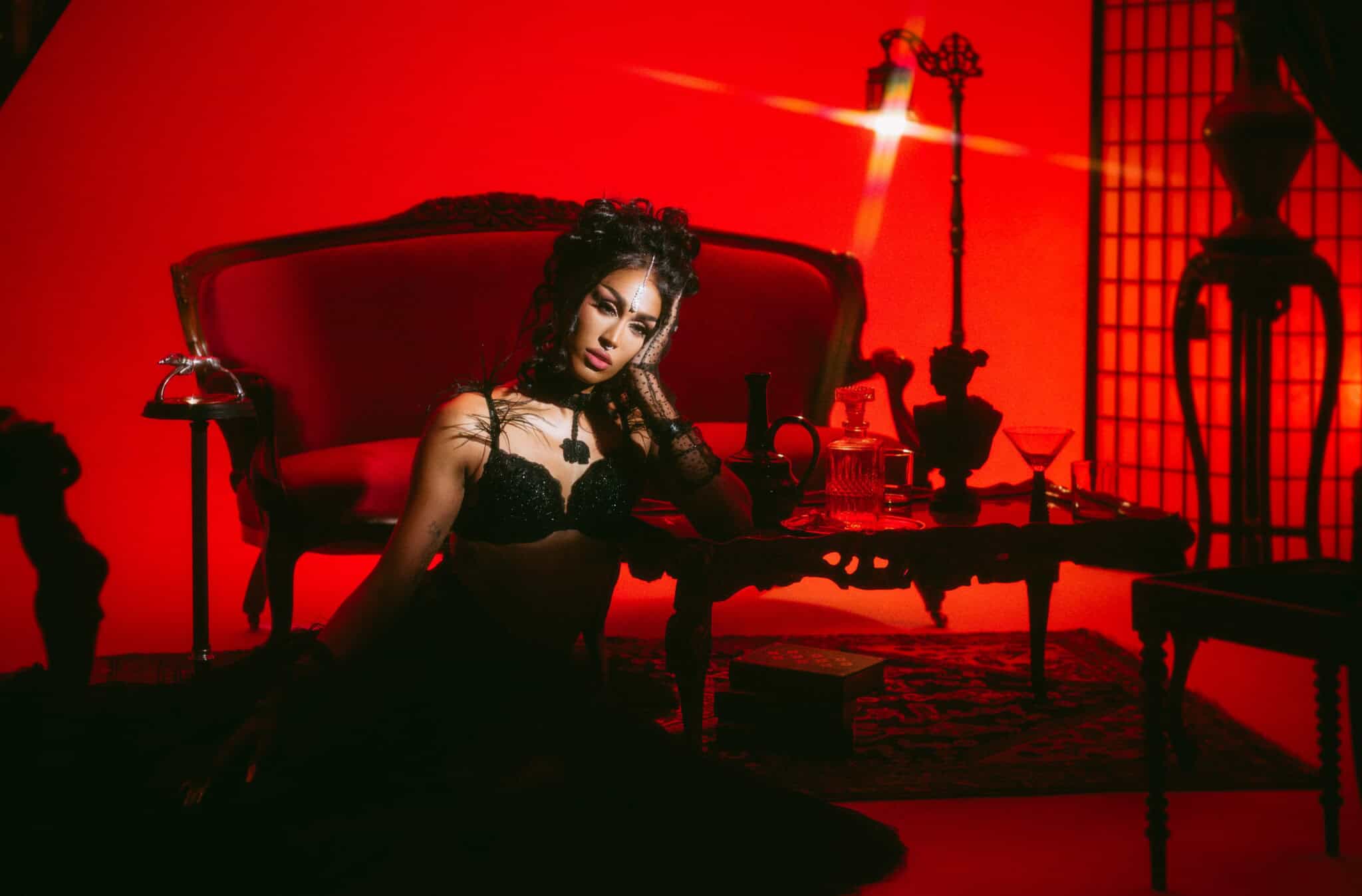A woman in black lingerie poses on the floor beside a vintage red sofa and ornate table, surrounded by gothic decor, in a room lit with dramatic red lighting.